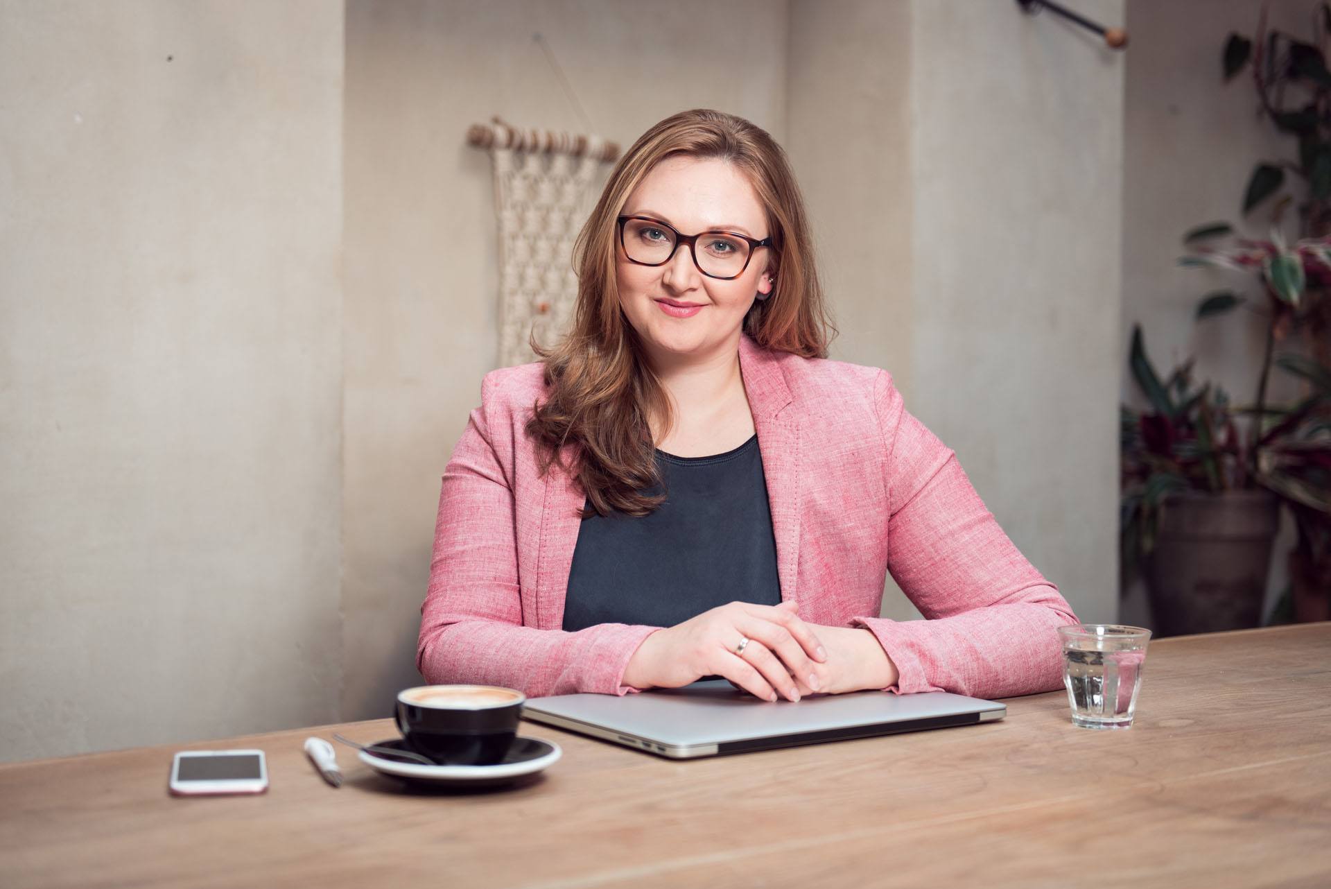 Eine junge Frau im rosa Blazer sitzt mit Laptop und Kaffeetasse in einem hippen Berliner Cafe und lächelt in die Kamera
